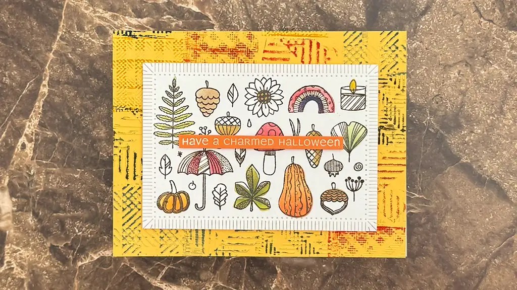 A handmade card on a marble surface showcases fall-themed drawings and the text "Have a Charmed Halloween" on an orange label—perfect for Autumn card ideas or inspiration for your Sept 25 club kits.