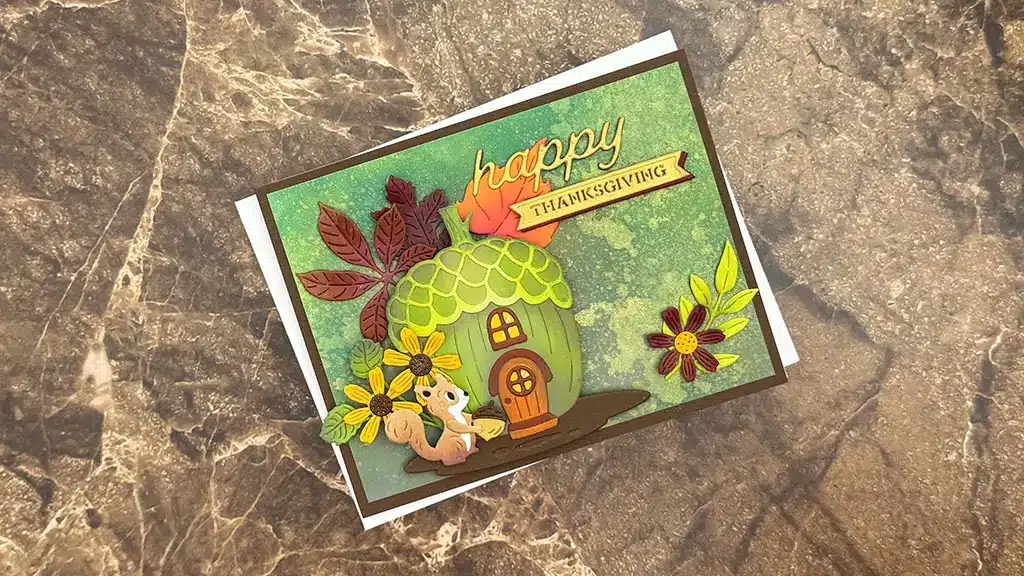 A handmade Thanksgiving card with a green acorn house, flowers, leaves, squirrels, and the words "happy THANKSGIVING" on a marbled surface. This card was created using Spellbinders' Sept 25 Club Kits Large Die of the month Acorn Acres.