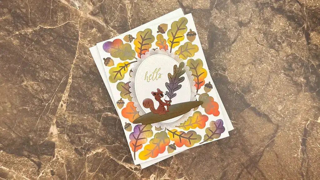 A greeting card with a squirrel holding a leaf, surrounded by colorful autumn leaves and acorns, on a brown marble surface—perfect inspiration for autumn card ideas or your next card making project. Thisi card is made using the Stencil Sept 25 Club kit.