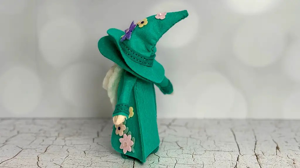 A small handmade 3D felt gnome figurine, wearing a green pointed hat and robe adorned with felt flowers, stands on a cracked white surface against a light blurred background.