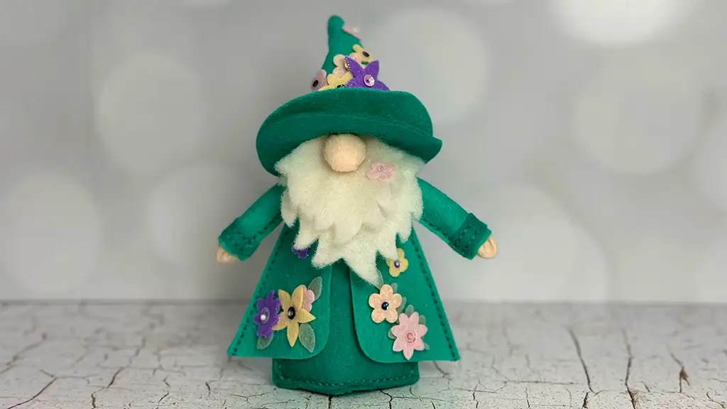 A handmade 3D Felt Gnome doll with a white beard, green hat, and coat decorated with colorful felt flowers stands on a cracked white surface.