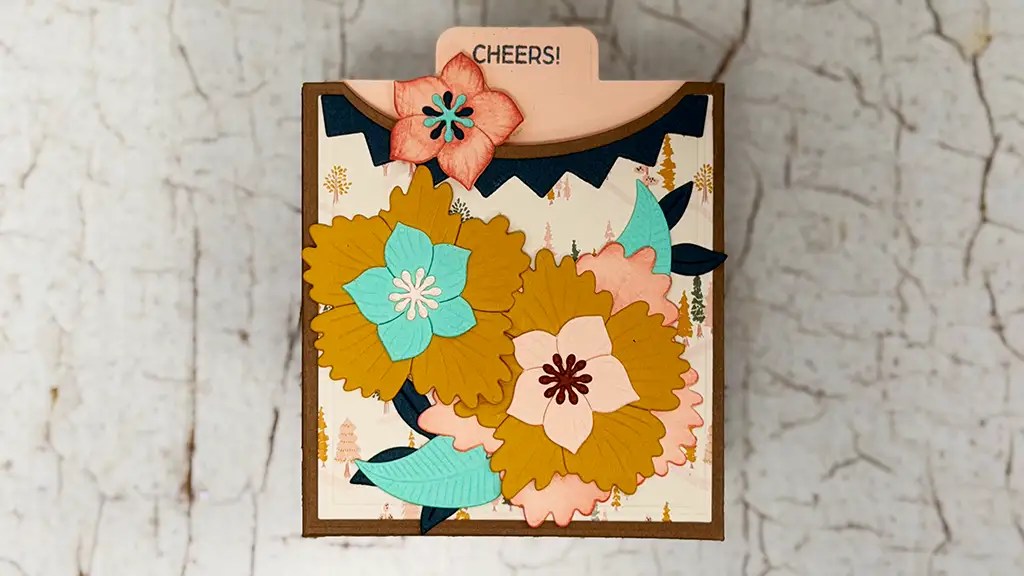 A handmade greeting card with layered paper flowers and "CHEERS!" on a tab at the top, set against a textured, cracked white background—perfect inspiration if you want to make birthday cards or try die cut birthday cards yourself.