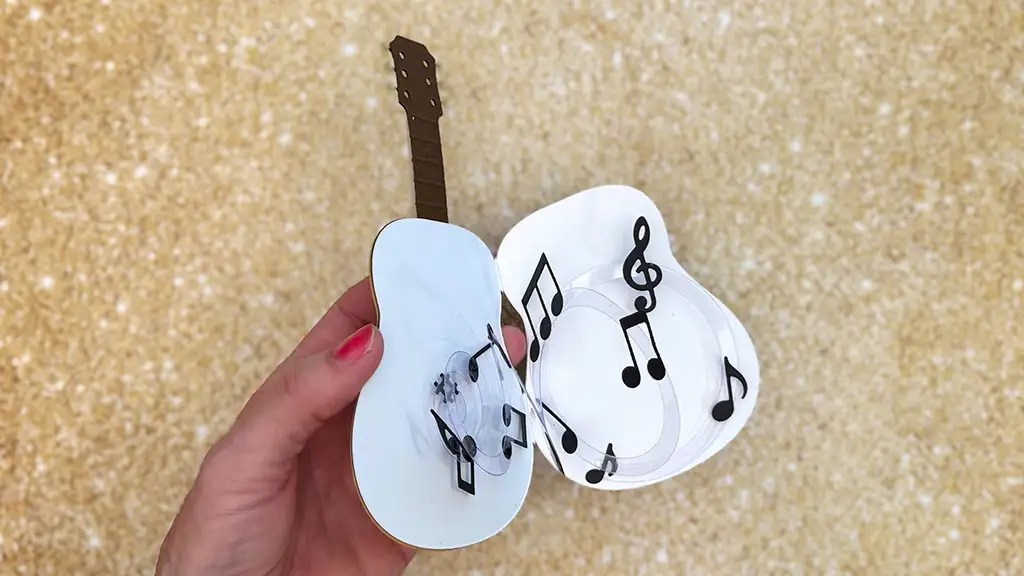 A hand holds an open card shaped like a guitar, revealing pop-up musical notes inside against a textured beige background.