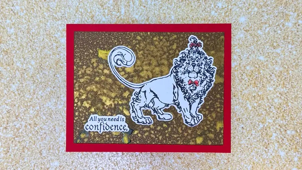 A handmade card with a lion drawing, red border, gold background, and the text "All you need is confidence. This card was created using products from Spellbinders Wonder of Oz Collection.