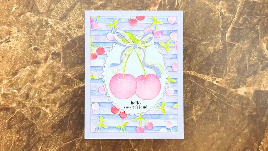 A greeting card from Spellbinders' Nov 2025 Kits featuring two large cherries tied with a ribbon and the text "hello sweet friend" on a colorful, cherry-themed background, placed on a brown marble surface.