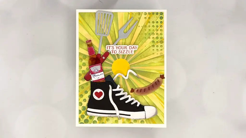 A greeting card for March 26 featuring a black sneaker, ketchup bottle, barbecue tools, sausage, and a sun with "IT'S YOUR DAY TO SIZZLE" on a yellow patterned background—perfect inspiration made with Spellbinders March 26 Club Kits or card making.