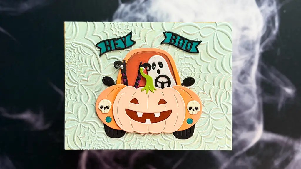 Halloween themed greeting card featuring a pink pumpkin car with skull accents and a ghost peeking from the trunk hey boo banner above on a embossed mint background