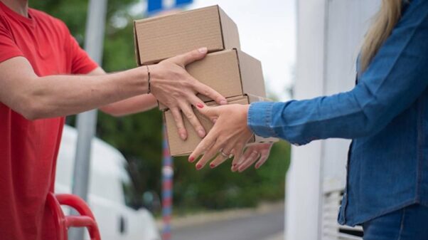 A red-shirted man handing a stack of three cardboard boxes to a woman in a blue shirt. Accessorial charges are fees for any additional services that the carrier has to complete during delivery.