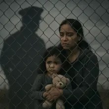 “Immigrant mother and child behind a chain-link fence with the shadow of a border agent looming over them.”