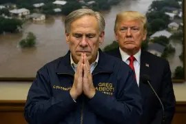 Texas Governor Greg Abbott stands with eyes closed and hands pressed in prayer, while former President Donald Trump looks on with a stern expression. Behind them, a blurred image of flooded homes and trees shows the devastation caused by flash floods in Texas.