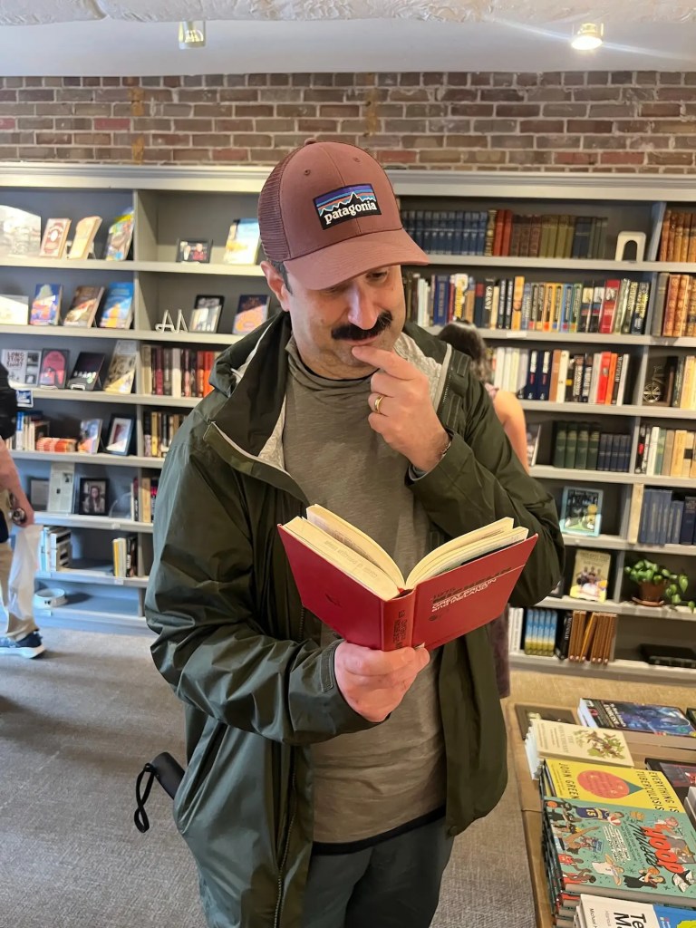 A man in a Patagonia cap and green jacket stands in Blue Bicycle Books, reading a small red book and playfully posing in thought, surrounded by shelves of novels. Alt-text generated with AI because the author is kinda lazy.