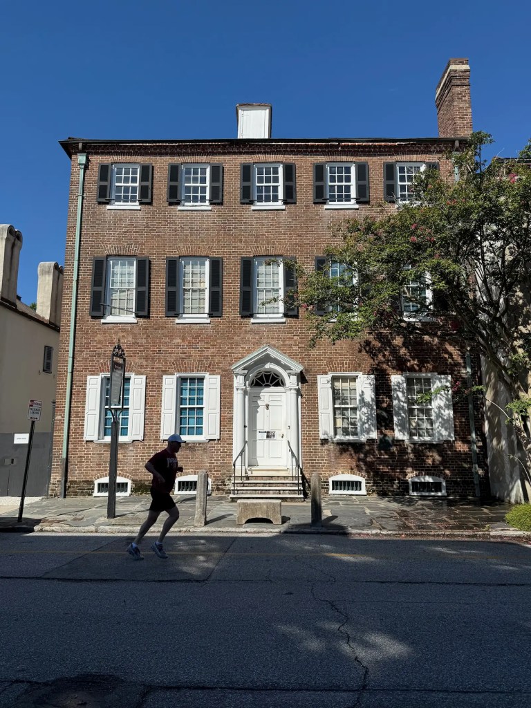 The Heyward-Washington House in Charleston, a Georgian brick home with black shutters and white trim, seen from across the street as a jogger passes by. Alt-text generated with AI because the author is kinda lazy.