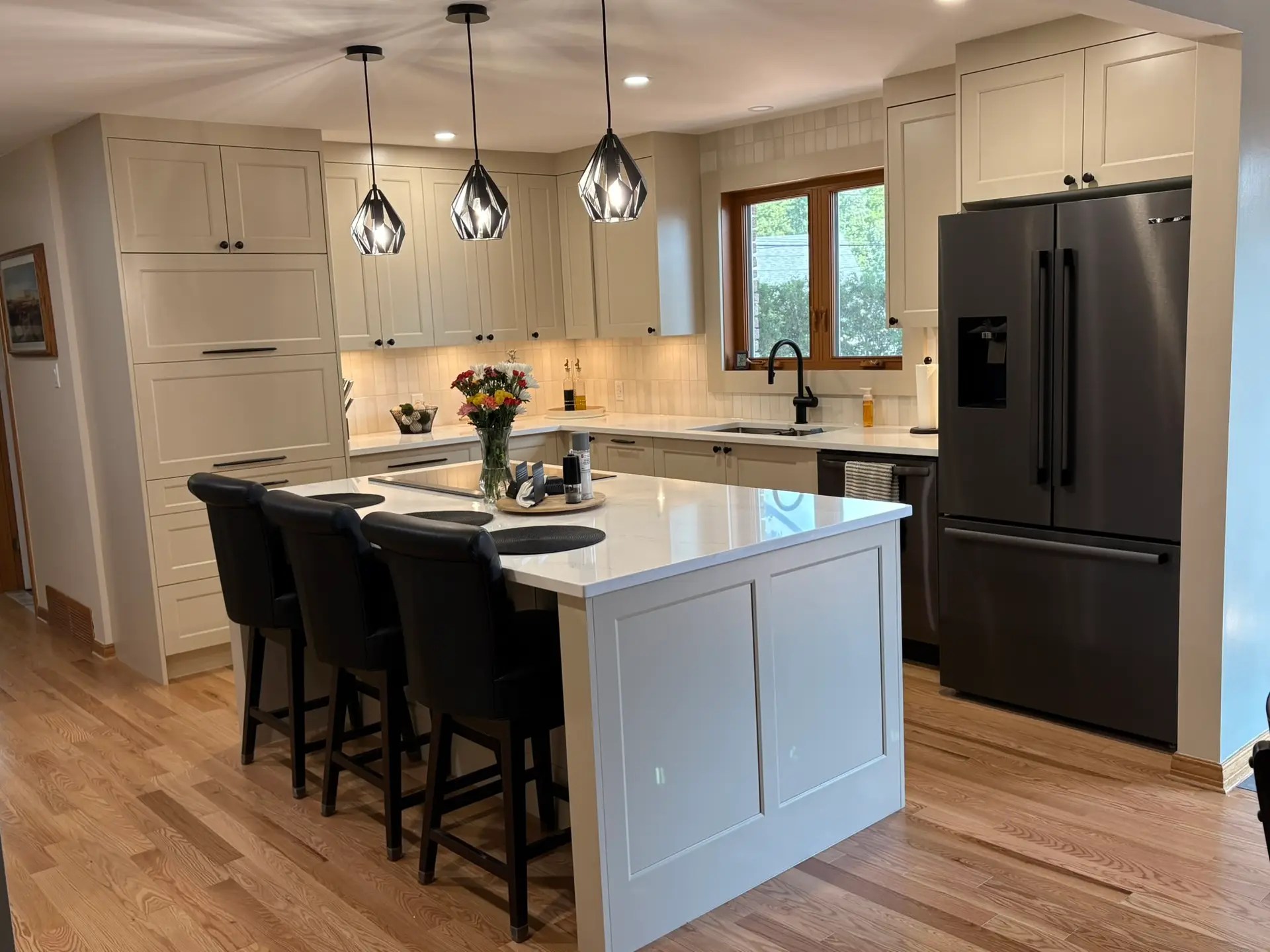 View back toward the large kitchen island with in island cooktop, downdraft venting, pendant lights, seating, and quartz countertop surrounded by latte toned cabinetry and engineered hardwood floors in a renovated Ottawa kitchen.
