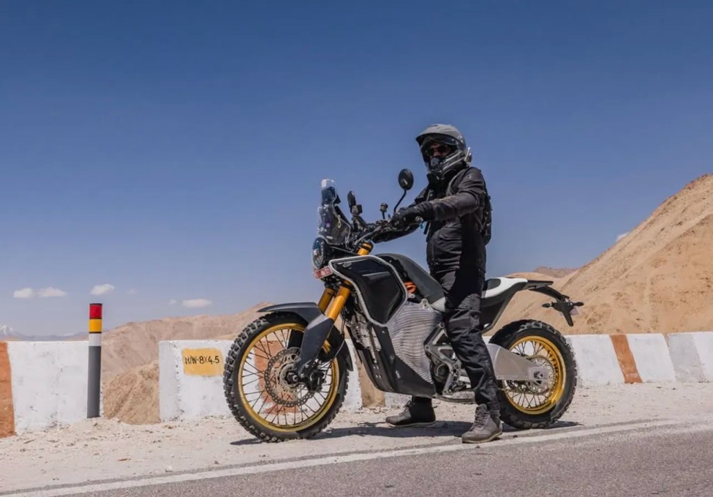 A rider in full gear stands next to a modern electric motorcycle on a road with mountains in the background, showcasing the bike's off-road capabilities.