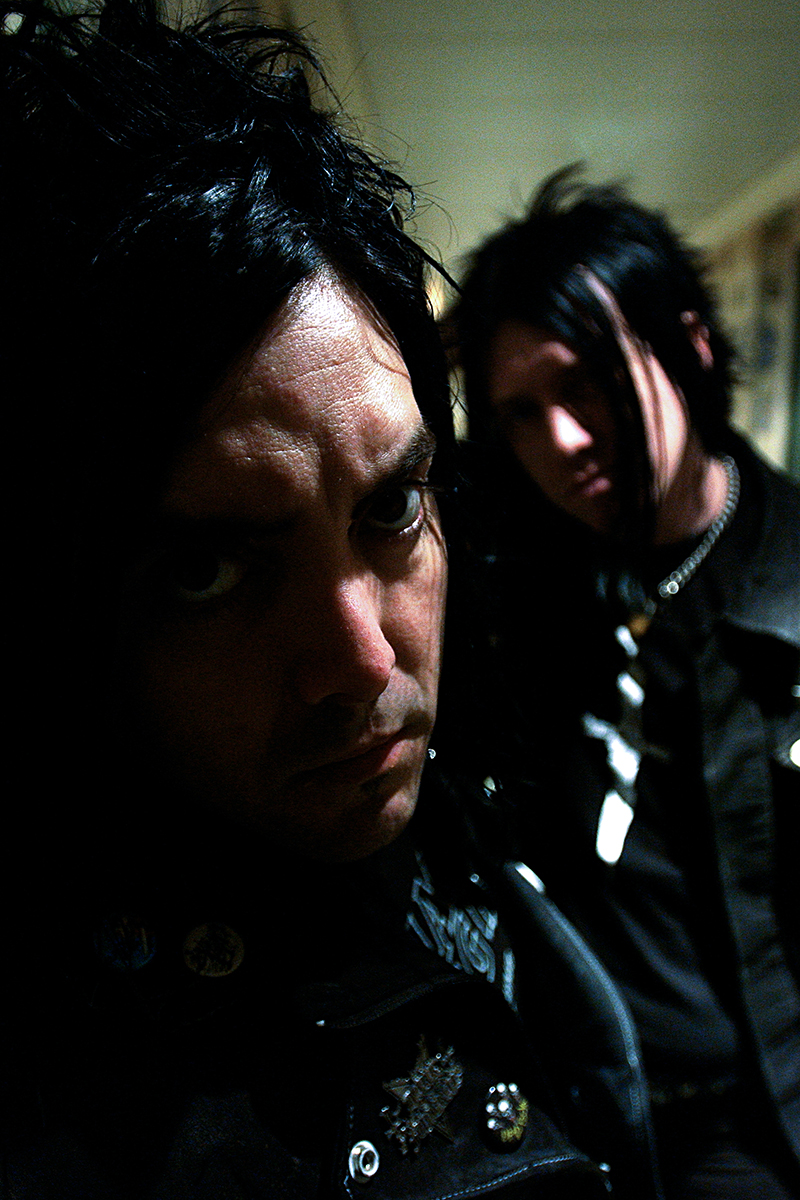 Portrait serré de Casey Chaos au premier plan, visage sombre et intense, avec un autre membre d’Amen flou en arrière-plan