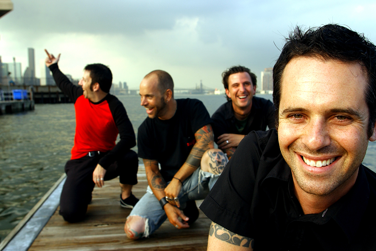 Portrait du groupe The Bouncing Souls sur un quai en bois, en bord de fleuve, souriants et détendus, avec la ville en arrière-plan