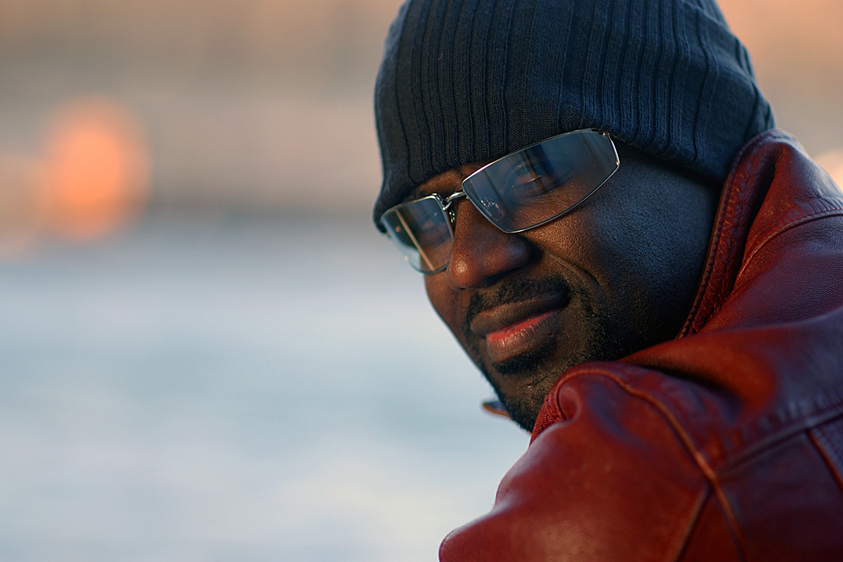 Portrait de Brian McKnight à Paris, portant un bonnet sombre, des lunettes et une veste en cuir rouge.
