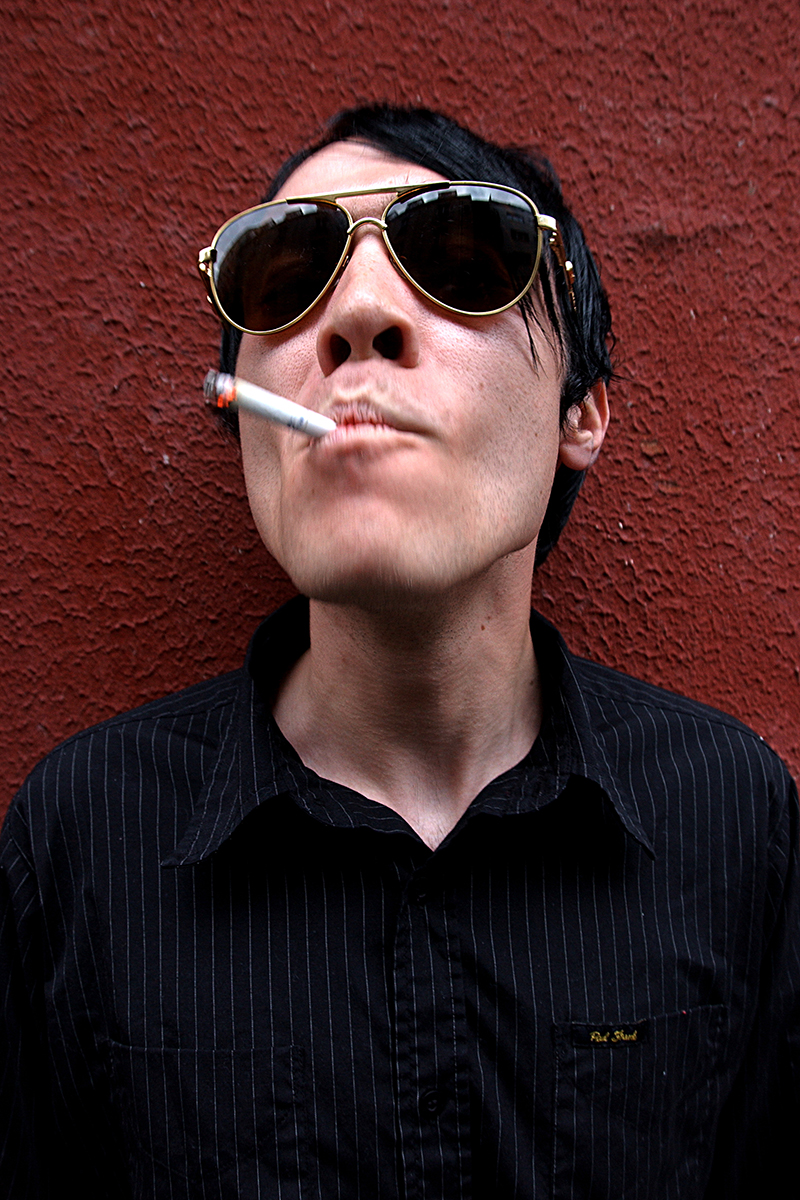 Portrait de Jorma Vik, batteur de The Bronx, cigarette aux lèvres et lunettes de soleil, devant un mur rouge.