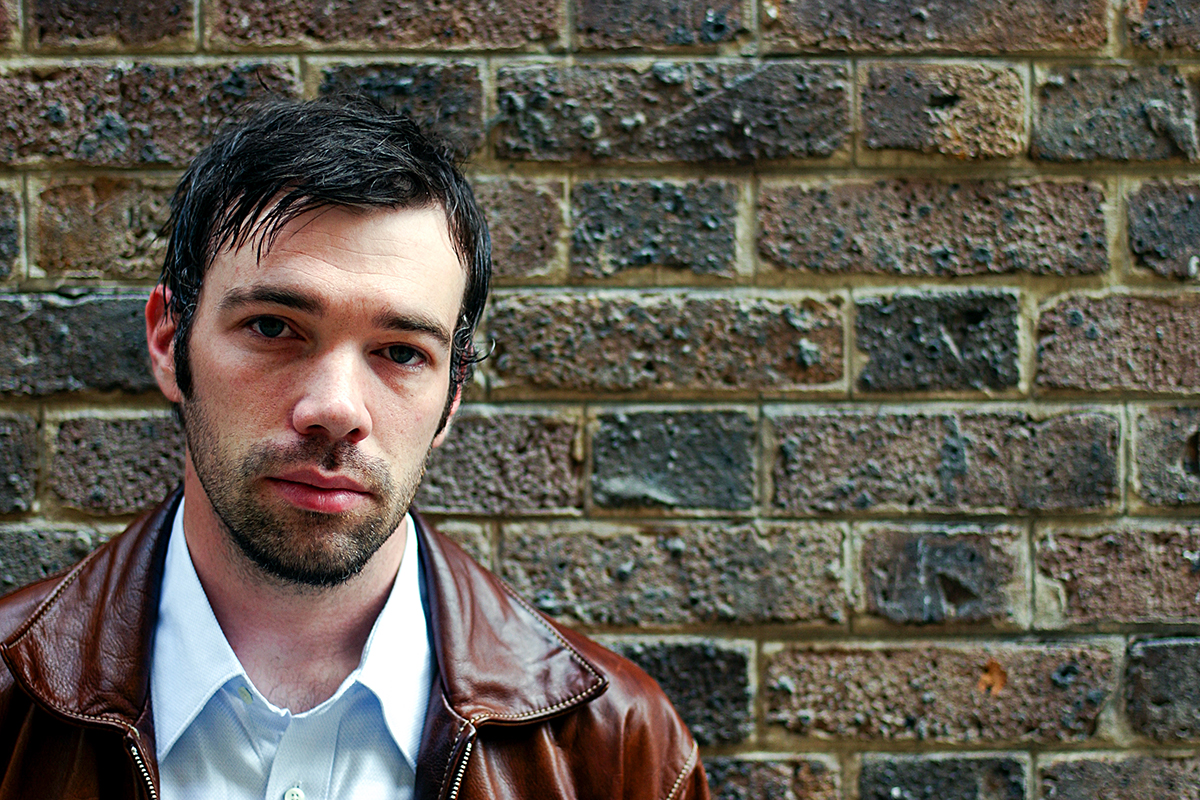 Portrait de Buck 65 devant un mur de briques, regard franc, veste en cuir et chemise claire