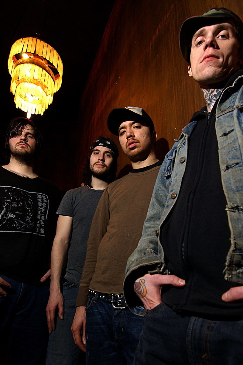 Les membres de Converge posent sous un lustre vintage, dans un couloir sombre aux murs cuivrés.