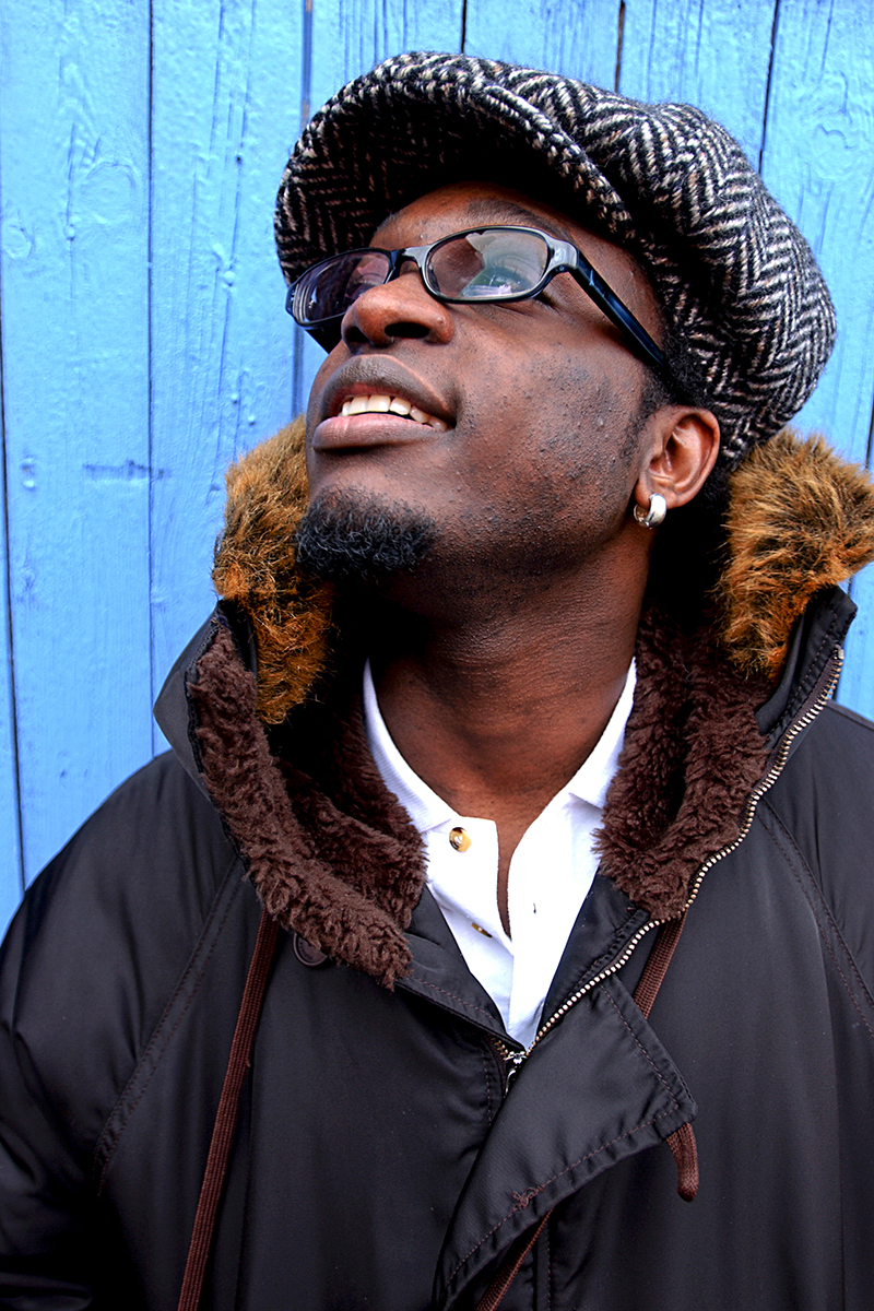 Portrait de Corneille en manteau à capuche et casquette en tweed, regardant vers le ciel devant une porte bleue