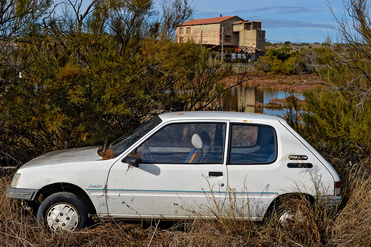 Voiture blanche abandonnée dans la nature près de Fitou, avec une maison en ruine en arrière-plan.