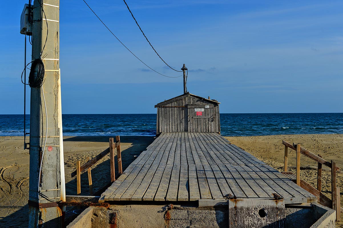 Jetée en bois menant à une cabane sur la plage de La Palme, avec la mer en arrière-plan.