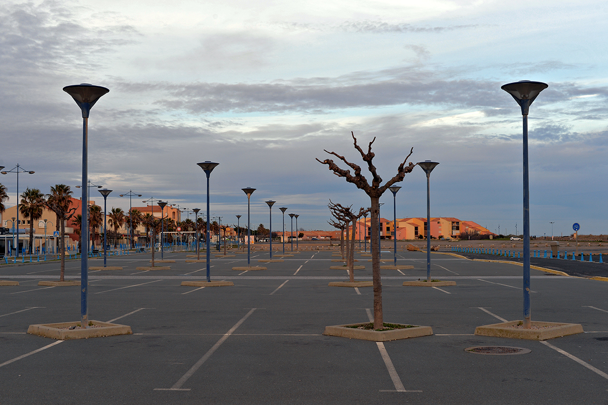 Parking vide à Saint-Pierre-la-Mer avec des lampadaires bleus et des arbres dénudés sous un ciel nuageux.