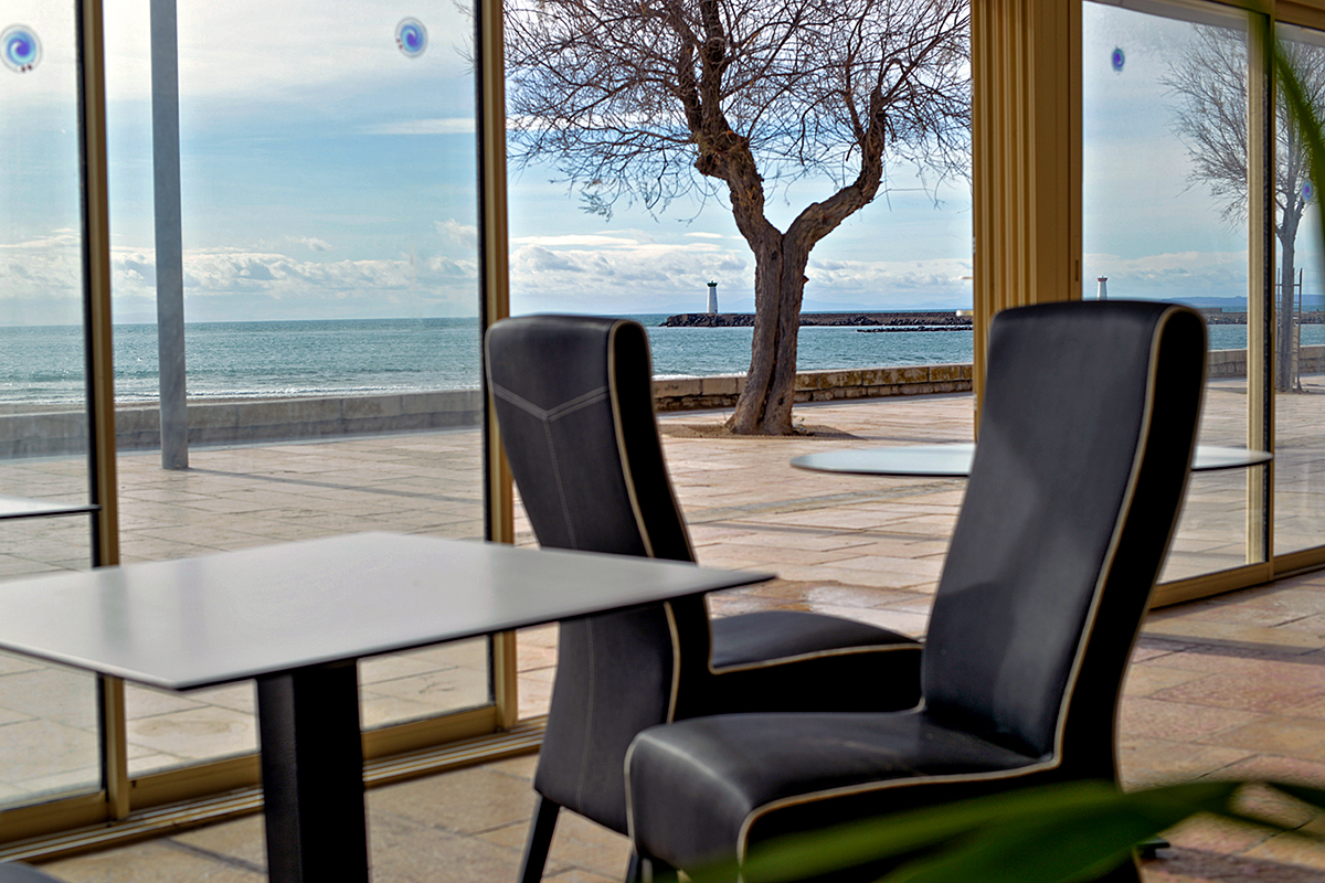 Vue depuis un intérieur au Grau d'Agde, avec un arbre et un phare au loin, surplombant la mer.