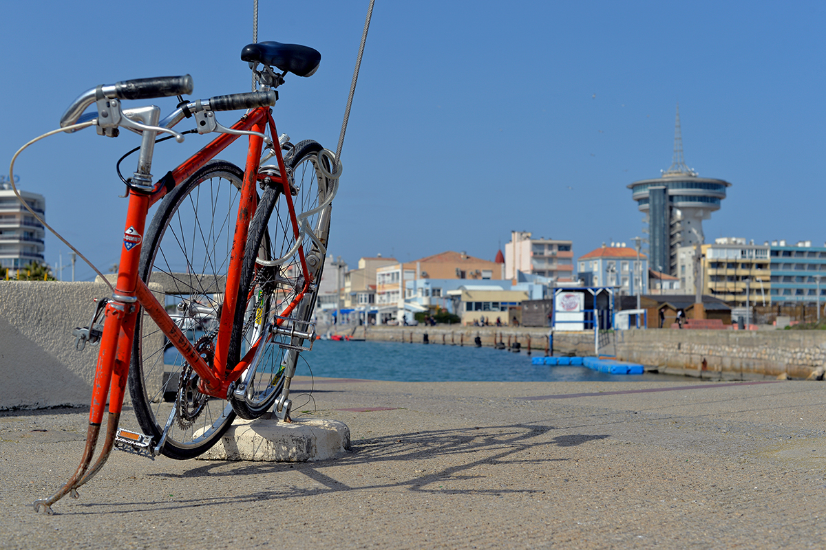 Vélo rouge à Palavas-les-Flots avec vue sur la ville et la mer en arrière-plan.