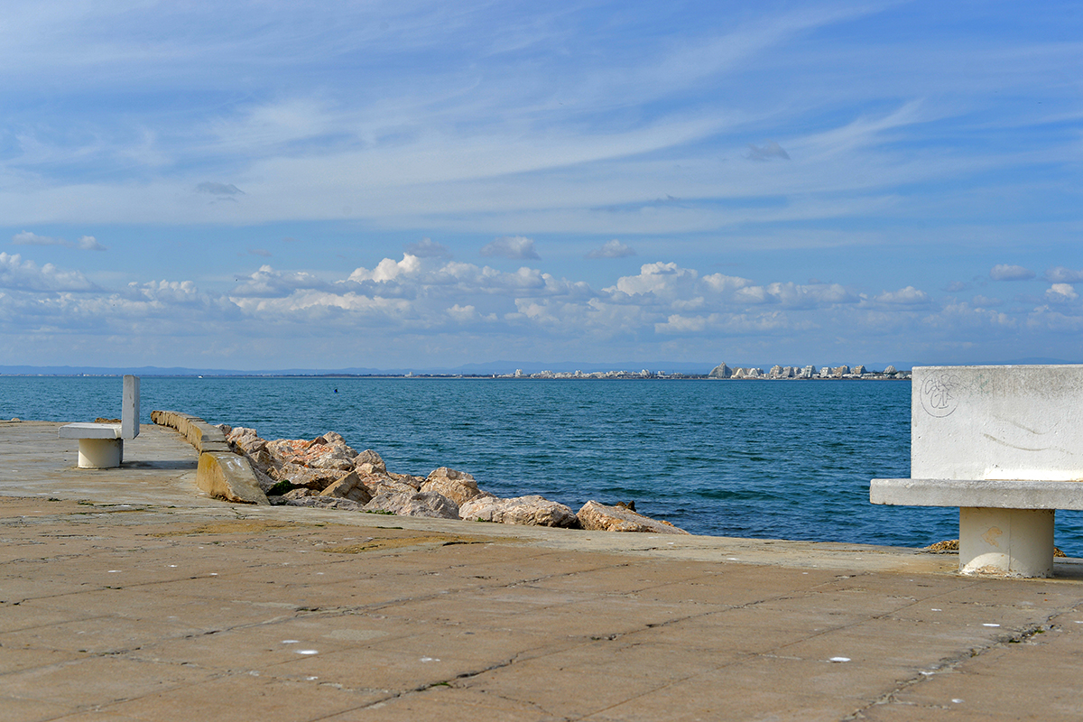 Vue sur la mer avec La Grande-Motte à l'horizon et un banc en béton au premier plan.