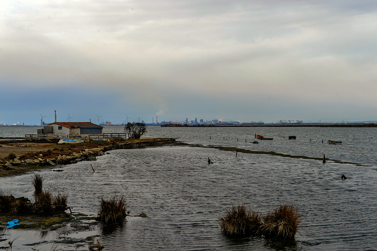 Vue du golfe de Fos-sur-Mer avec une maison au bord de l'eau et des installations industrielles à l'horizon.