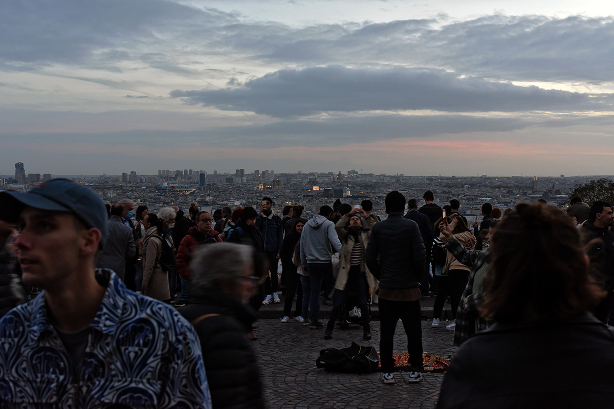 Vue panoramique de Paris au crépuscule depuis le parvis du Sacré-Cœur à Montmartre, avec une foule de passants et de touristes profitant du spectacle urbain.