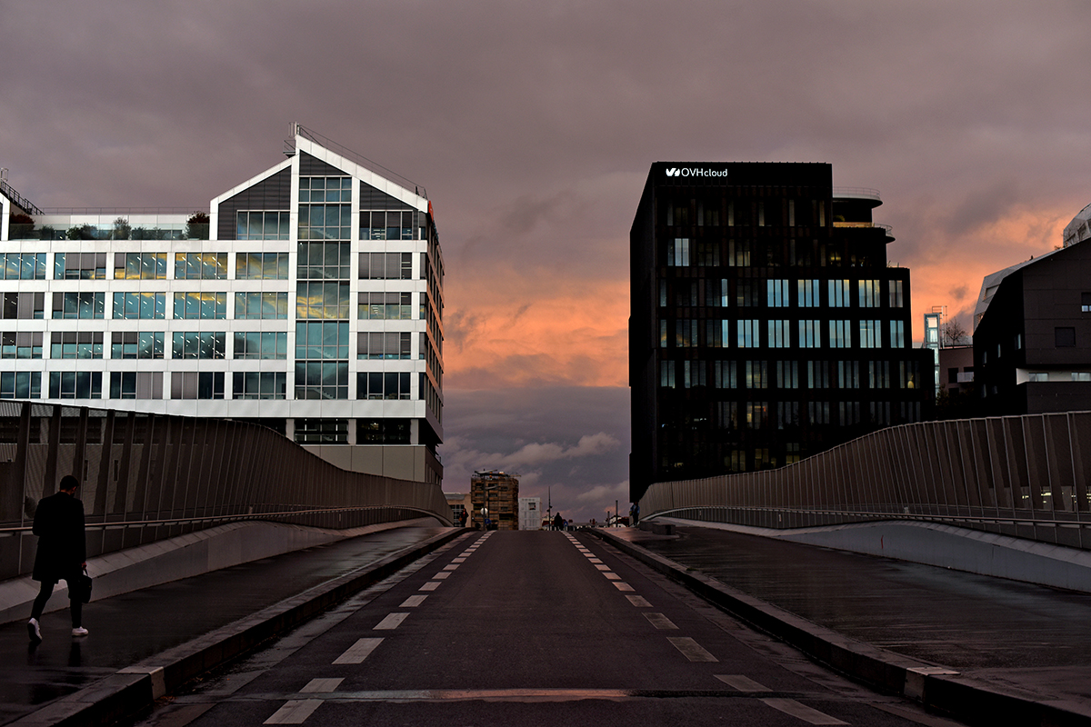 Vue urbaine du quartier en développement du 17e arrondissement de Paris, rue Mère Teresa, au crépuscule. Une passerelle encadrée par des immeubles modernes sous un ciel orageux teinté de lumière.