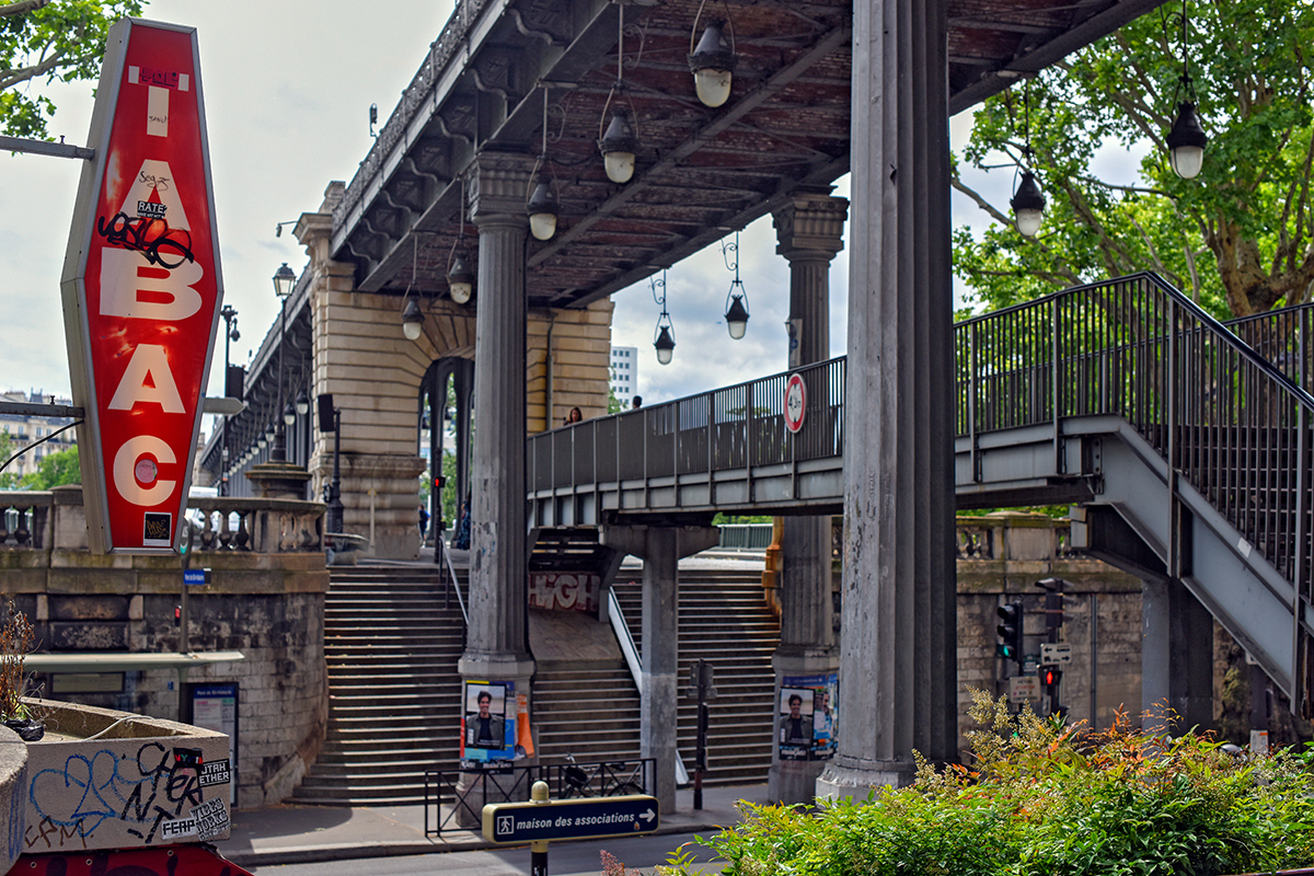 Passerelle piétonne sous un viaduc du métro aérien parisien, avec escaliers, poteaux en fonte et enseigne rouge de bar.