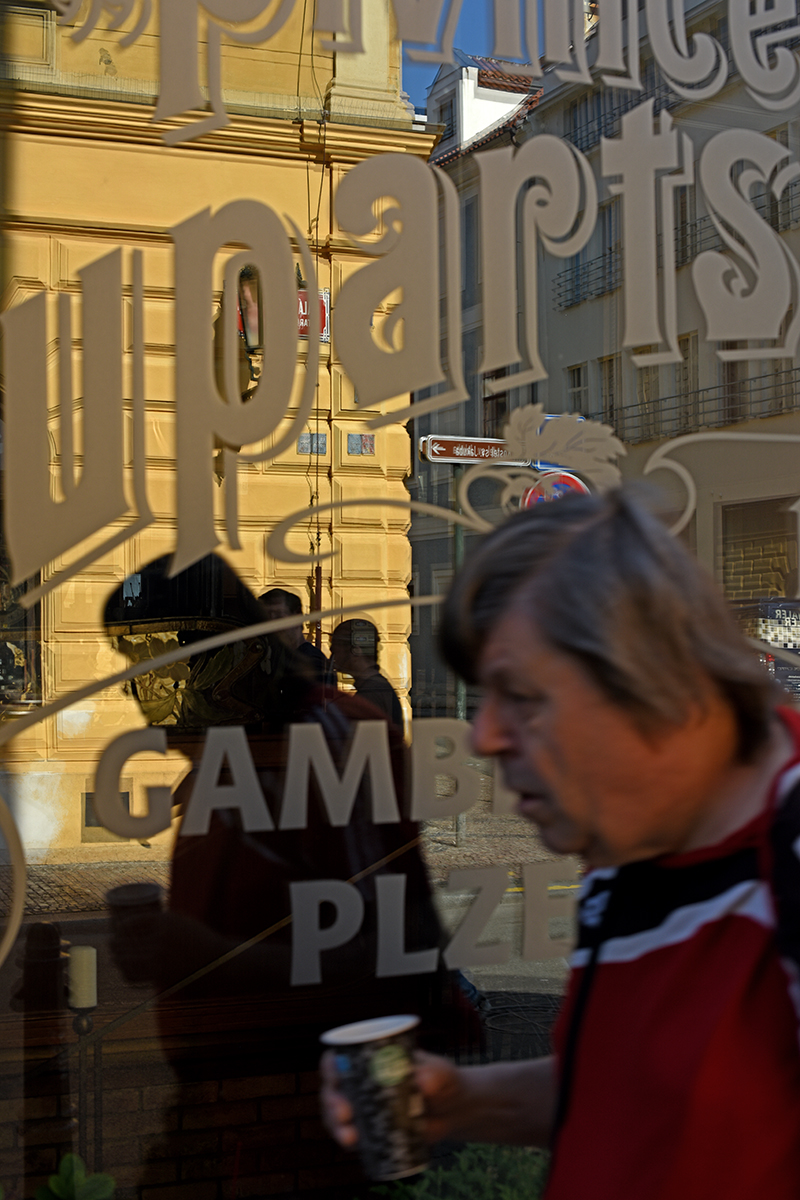 Reflets d'un bâtiment et d'une enseigne "Gambrinus Plzeň" dans le centre de Prague, avec une personne en premier plan.