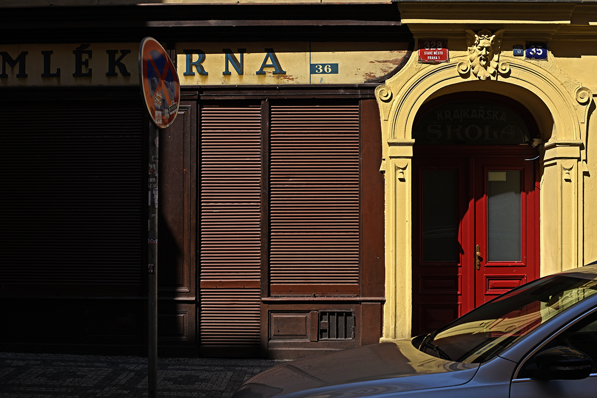 Façade d'un bâtiment dans le centre de Prague avec des volets fermés et une porte rouge, sous une enseigne "MLEKÁRNA".