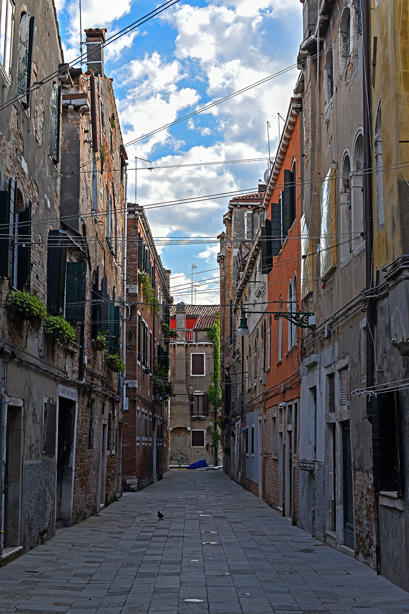 Ruelle étroite et pavée de Venise, bordée de bâtiments anciens et usés sous un ciel bleu.