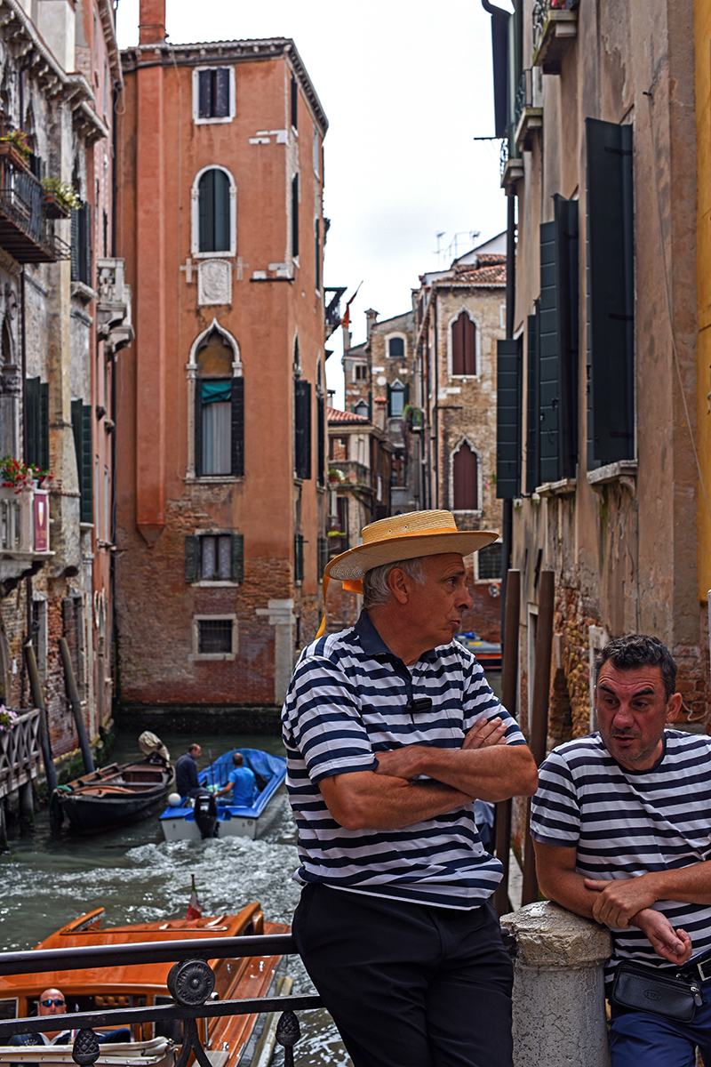 Deux gondoliers en tenue traditionnelle discutant près d'un canal à Venise, avec des gondoles en arrière-plan.