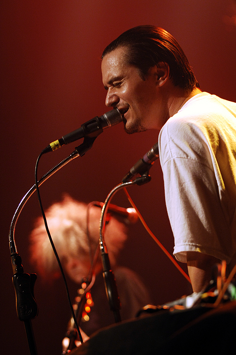 Mike Patton chantant sur scène avec Fantômas, sous une lumière rouge, accompagné d’un autre musicien en arrière-plan.