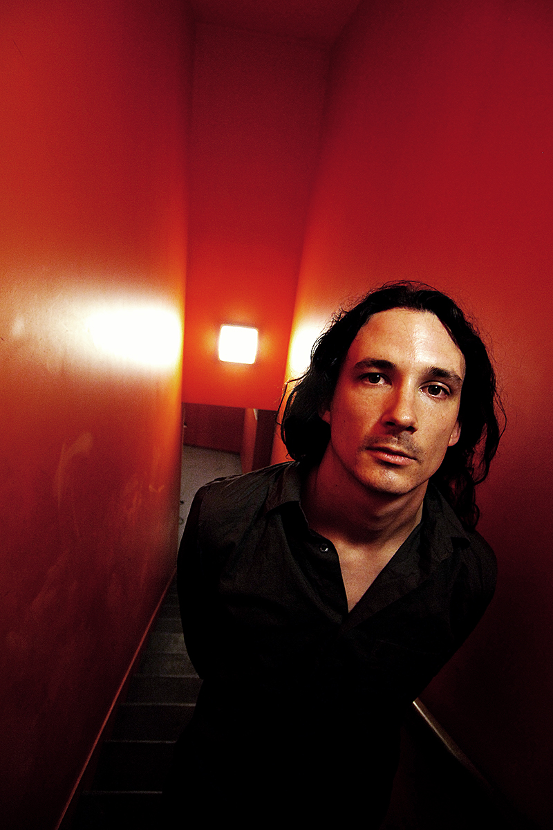 Joe Duplantier, chanteur et guitariste du groupe Gojira, photographié dans un escalier aux murs rouges. © Daniel Mielniczek.