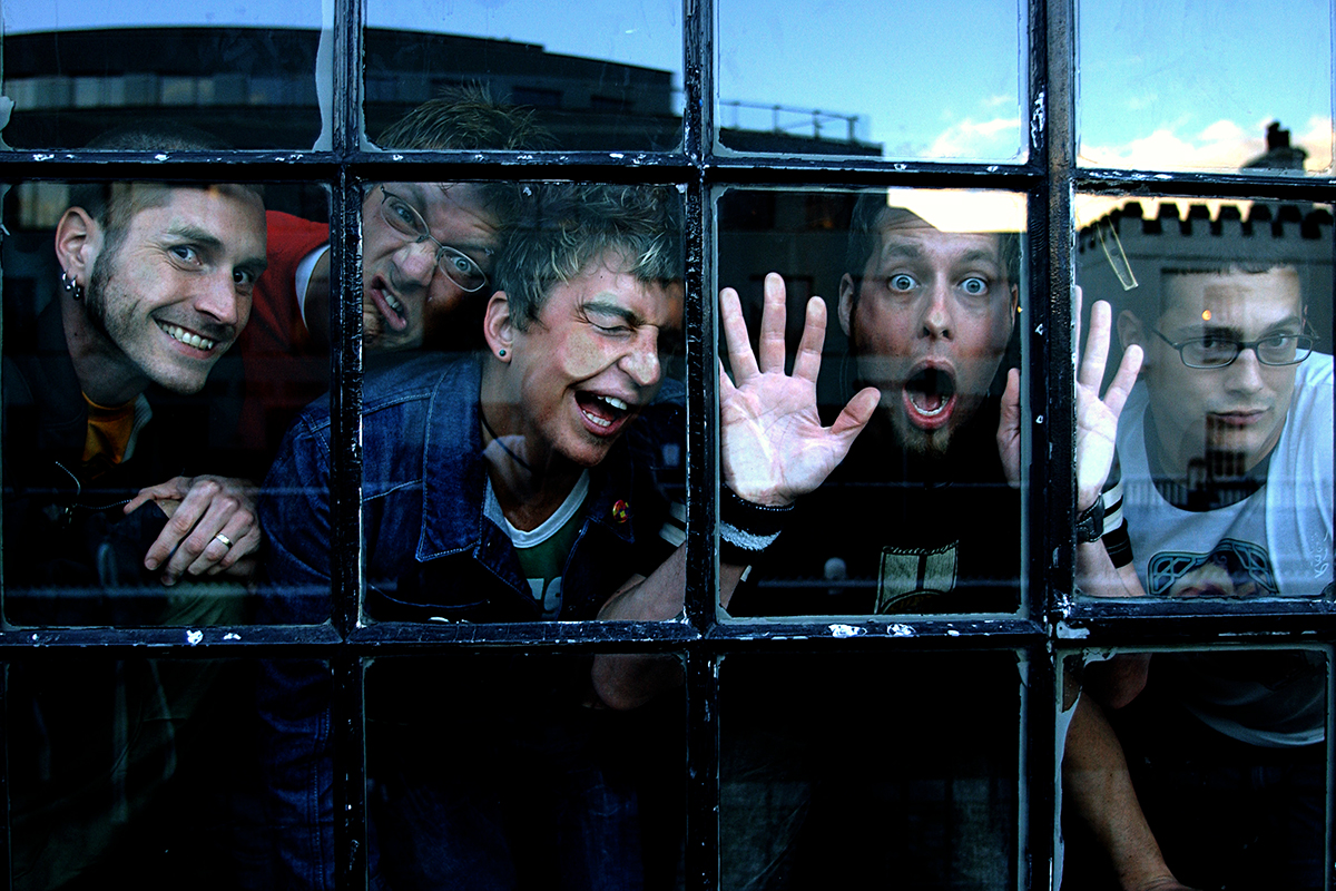 Le groupe Hot Action Cop vu à travers une fenêtre à petits carreaux, faisant des grimaces et poses expressives.