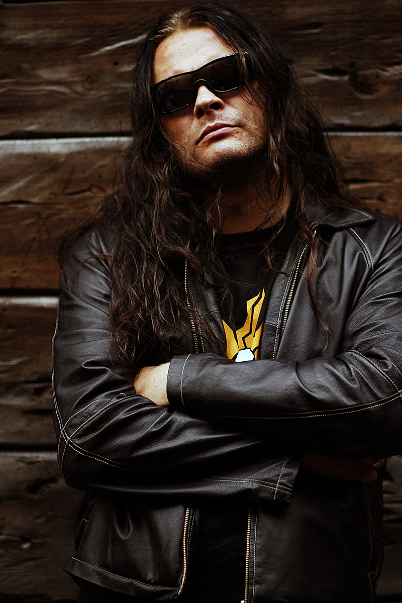 Abbath, musicien de black metal norvégien, en veste de cuir et lunettes noires, bras croisés devant un mur en bois