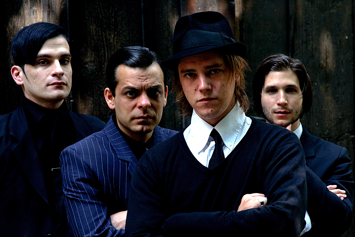 Portrait du groupe Interpol en costume sombre, l’air grave, posant devant une paroi boisée