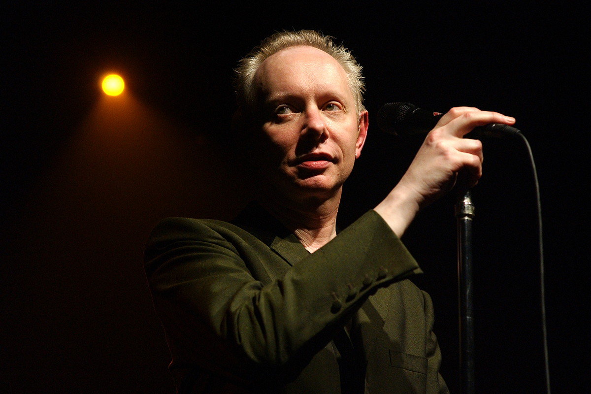 Joe Jackson sur scène à Paris, tenant un micro sous une lumière tamisée.