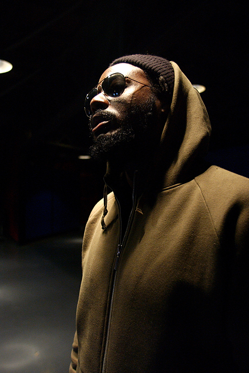 Portrait de K-OS, en sweat à capuche et lunettes de soleil, éclairé par le haut