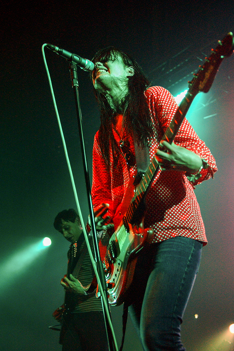 Alison Mosshart du groupe The Kills sur scène, en train de chanter et jouer de la guitare, éclairée par des lumières rouges et vertes