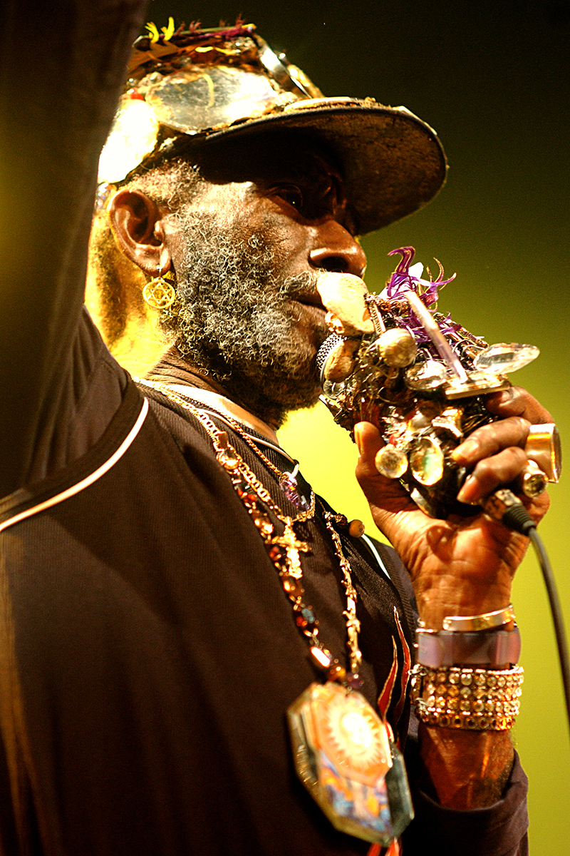 Lee "Scratch" Perry en concert à l’Élysée Montmartre, Paris, micro orné d’objets symboliques, ambiance dorée.