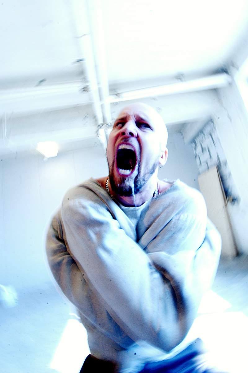 Portrait expressif de Jens Kidman, chanteur de Meshuggah, hurlant en camisole de force dans une pièce blanche surexposée