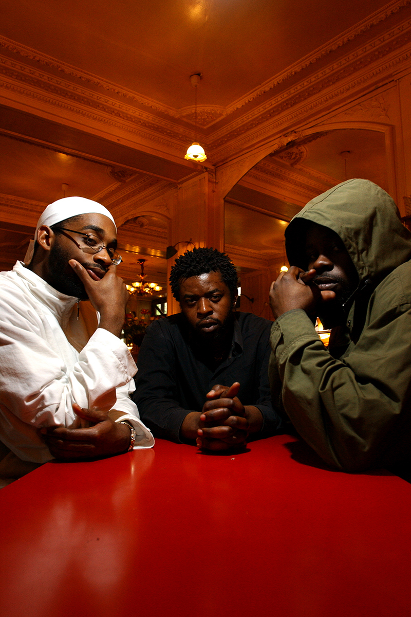 Portrait du groupe de rap français OFX, réuni autour d’une table rouge dans un intérieur au décor classique